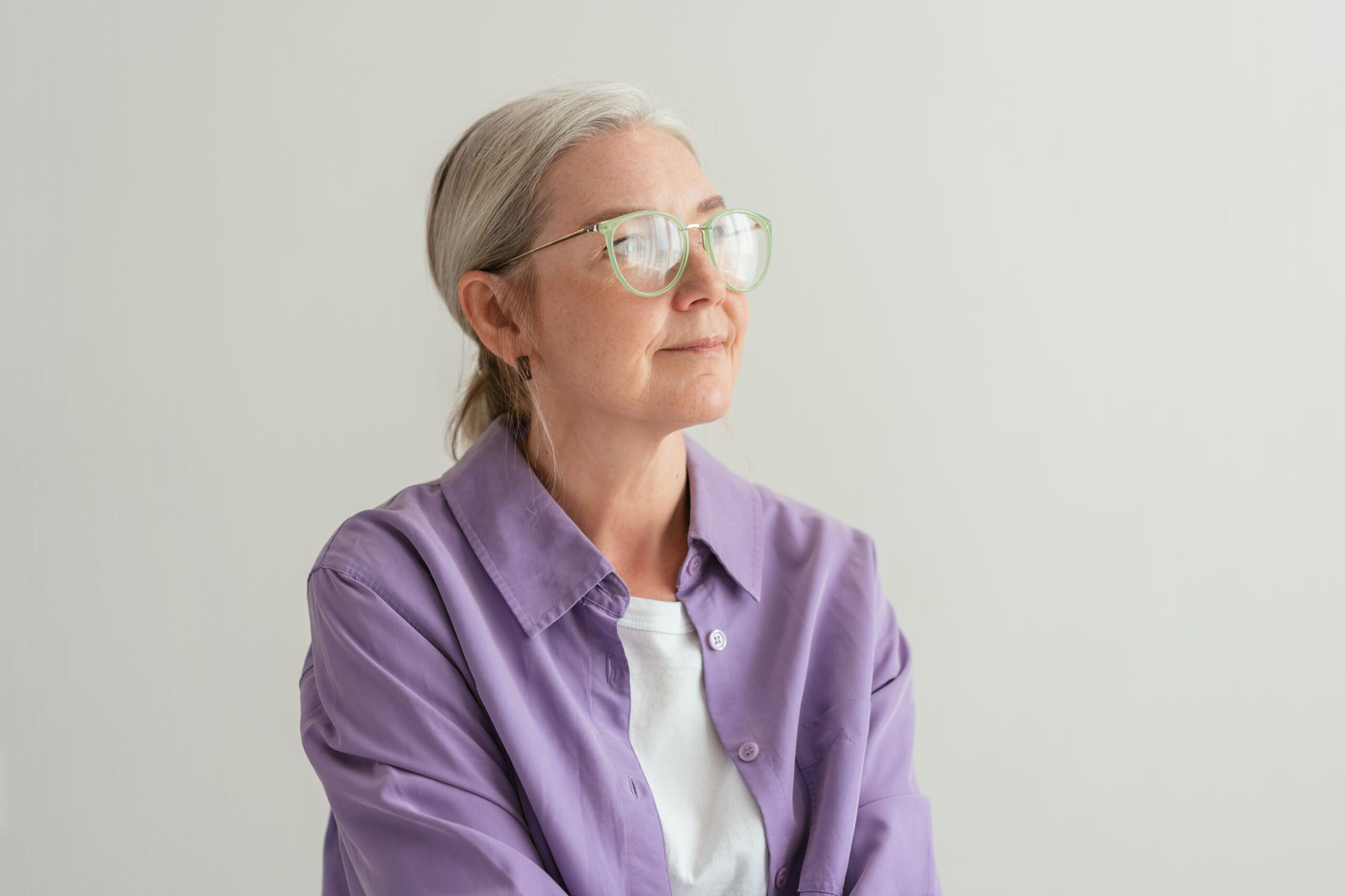 Home Senior woman with grey hair wearing eyeglasses and a purple shirt, looking thoughtful.